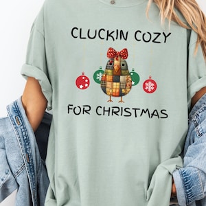 以下が含まれることがあります： 「CLUCKIN COZY FOR CHRISTMAS」の文字が入った、薄い緑色のTシャツ。赤いリボン、吊り下げられたオーナメント、雪の結晶のオーナメントが付いた、パッチワークの鶏のデザインが特徴です。