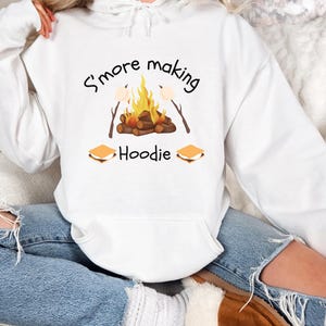 May include: White hoodie with a campfire and s'mores graphic. The text on the hoodie reads "S'more making Hoodie".