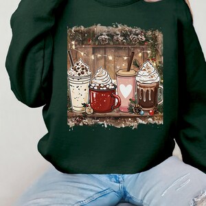 Peut inclure: Sweat-shirt vert fonc&eacute; avec un motif de quatre boissons chaudes festives : une boisson blanche avec des p&eacute;pites de chocolat, une tasse rouge avec des guimauves, une tasse rose avec un c&oelig;ur et une tasse marron avec de la cr&egrave;me fouett&eacute;e.