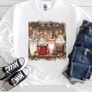 Peut inclure: Sweat-shirt blanc avec un motif festif de quatre boissons chaudes : un milkshake, une tasse de chocolat chaud, une tasse rose avec un c&oelig;ur et une tasse de chocolat chaud, le tout garni de cr&egrave;me fouett&eacute;e. Le motif est plac&eacute; sur un fond en bois.