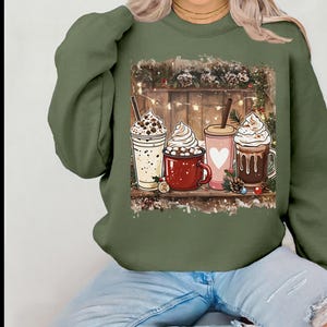 Peut inclure: Un sweat-shirt vert olive avec un motif de quatre boissons festives : un milk-shake, une tasse de chocolat chaud, une tasse rose avec un c&oelig;ur et une tasse de caf&eacute;. Les boissons sont d&eacute;cor&eacute;es de cr&egrave;me fouett&eacute;e et d'autres garnitures.
