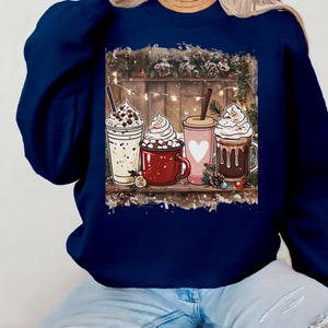 Peut inclure: Sweat-shirt bleu marine avec un motif de quatre boissons chaudes festives, chacune garnie de cr&egrave;me fouett&eacute;e. L'image comprend une &eacute;tag&egrave;re en bois et des d&eacute;corations de No&euml;l. Un style d&eacute;contract&eacute; et confortable.