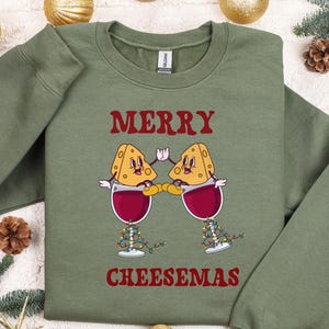 May include: Green sweatshirt with a red graphic of two cheese wedges holding glasses of wine. The text "Merry Cheesemas" is printed below the graphic.
