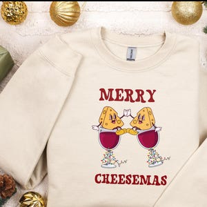 May include: A beige sweatshirt with the text "Merry Cheesemas" in red. Two cartoon cheese wedges are holding glasses of red wine.