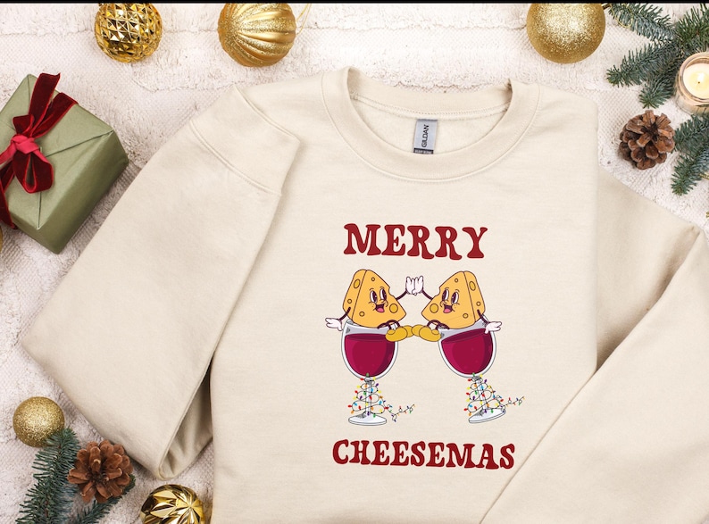 May include: A beige sweatshirt with the text "Merry Cheesemas" in red. Two cartoon cheese wedges are holding glasses of red wine.