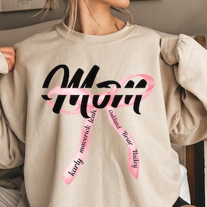 Puede incluir: Una sudadera beige con un gráfico negro de "Mom". La palabra "Mom" está envuelta en una cinta rosa con los nombres "Karly", "Maverick", "Leah", "Oakland", "Briar" y "Bailey" escritos en la cinta.