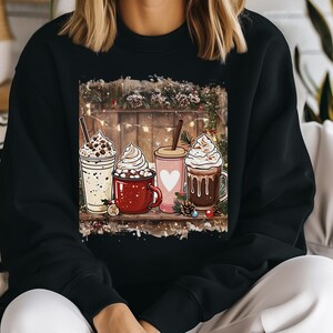 Peut inclure: Sweatshirt noir avec une illustration festive de quatre boissons chaudes. L'image montre une boisson blanche avec des p&eacute;pites de chocolat, une tasse rouge avec des guimauves, une tasse rose avec un c&oelig;ur et une tasse marron avec du chocolat. Le fond a une cl&ocirc;ture en bois et des d&eacute;corations de No&euml;l.