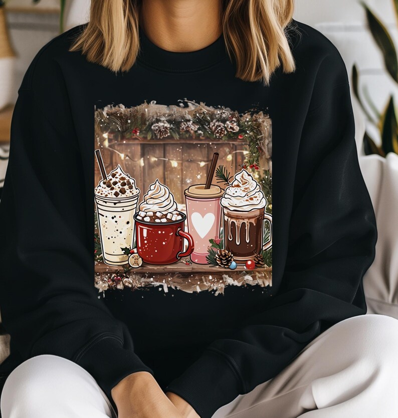 Peut inclure: Sweatshirt noir avec une illustration festive de quatre boissons chaudes. L'image montre une boisson blanche avec des p&eacute;pites de chocolat, une tasse rouge avec des guimauves, une tasse rose avec un c&oelig;ur et une tasse marron avec du chocolat. Le fond a une cl&ocirc;ture en bois et des d&eacute;corations de No&euml;l.