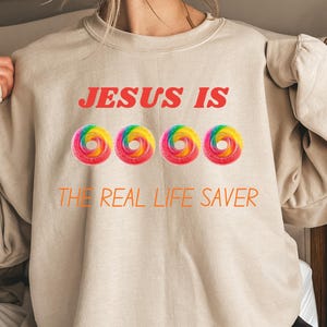 May include: Beige sweatshirt with the text "JESUS IS" in red above four colorful ring-shaped designs, and "THE REAL LIFE SAVER" in orange below. The sweatshirt is a casual, comfortable style.