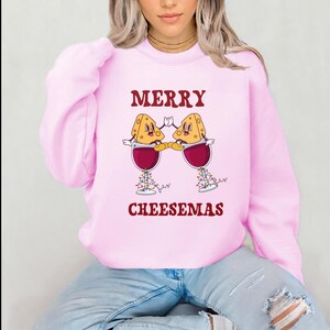 May include: Pink sweatshirt with a festive design featuring two cartoon cheese wedges holding glasses of wine with Christmas lights. The text "Merry Cheesemas" is printed below the design.