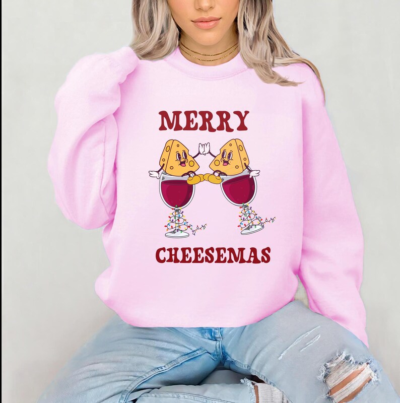 May include: Pink sweatshirt with a festive design featuring two cartoon cheese wedges holding glasses of wine with Christmas lights. The text "Merry Cheesemas" is printed below the design.