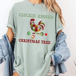 以下が含まれることがあります： 「CLUCKIN AROUND CHRISTMAS TREE」の文字が入った、薄い緑色のTシャツ。カラフルなニワトリのグラフィックとクリスマスオーナメントが特徴です。お祭り気分を盛り上げるカジュアルウェアです。