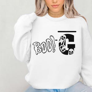 Pode incluir: Camisola branca com a frase "Boo!" em preto e um "G" estilizado com ilustrações de caveiras. Uma peça de vestuário casual de manga comprida.