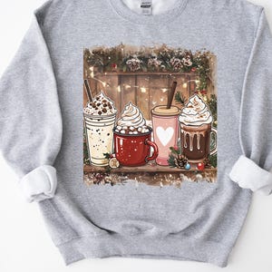 Peut inclure: Un sweat-shirt gris clair avec un motif de quatre boissons chaudes : un milkshake, une tasse de chocolat chaud, une tasse rose avec un c&oelig;ur et un caf&eacute;. L'arri&egrave;re-plan comprend une cl&ocirc;ture en bois, des guirlandes lumineuses et des d&eacute;corations festives.