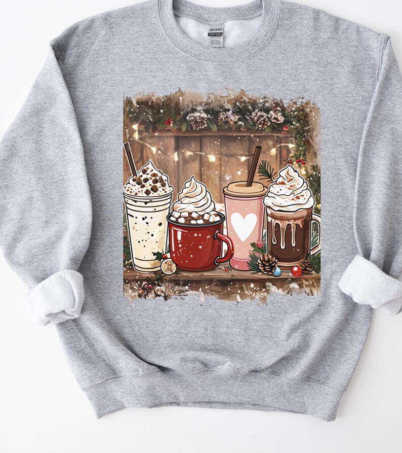 Peut inclure: Un sweat-shirt gris clair avec un motif de quatre boissons chaudes : un milkshake, une tasse de chocolat chaud, une tasse rose avec un c&oelig;ur et un caf&eacute;. L'arri&egrave;re-plan comprend une cl&ocirc;ture en bois, des guirlandes lumineuses et des d&eacute;corations festives.