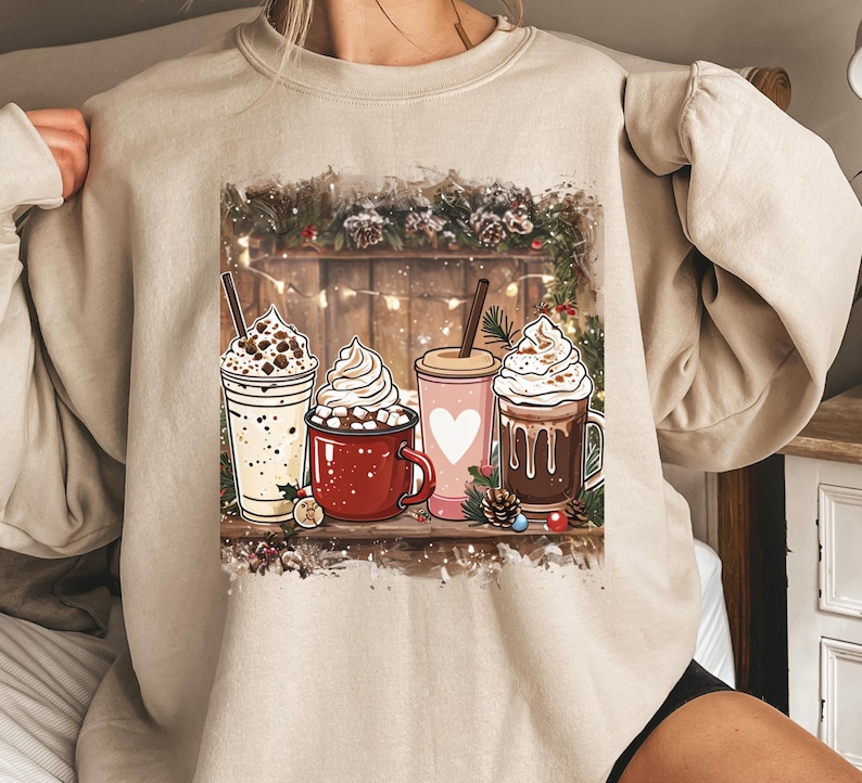 Peut inclure: Sweat-shirt beige avec un motif de quatre boissons festives : un caf&eacute; glac&eacute; &agrave; la vanille, une tasse rouge de chocolat chaud, une boisson rose avec un c&oelig;ur et une boisson au chocolat. Chaque boisson est garnie de cr&egrave;me fouett&eacute;e. Le fond comprend des planches de bois et des d&eacute;corations de vacances.