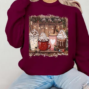 Peut inclure: Un sweat-shirt bordeaux avec un motif de quatre boissons chaudes festives &agrave; la cr&egrave;me fouett&eacute;e. Les boissons sont dans diverses tasses et mugs, sur fond de bois avec des d&eacute;corations de No&euml;l. Le sweat-shirt est port&eacute; par une personne en jean bleu.