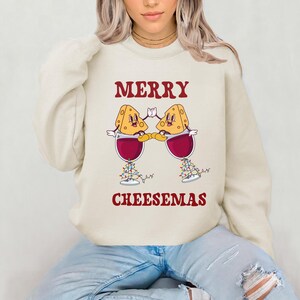 May include: A cream colored sweatshirt with the text "Merry Cheesemas" in red. The design features two cartoon cheese wedges holding glasses of red wine.