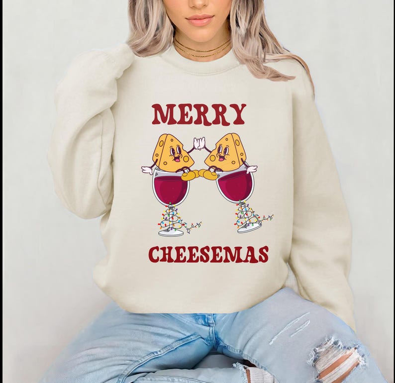 May include: A cream colored sweatshirt with the text "Merry Cheesemas" in red. The design features two cartoon cheese wedges holding glasses of red wine.