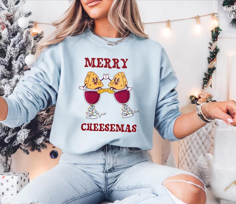 May include: A light blue sweatshirt with the text "Merry Cheesemas" in red. The design features two cartoon cheese wedges wearing Santa hats and holding glasses of wine.