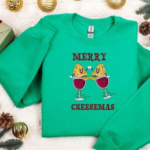 May include: Green Christmas sweater with a festive design featuring two cheese wedges wearing Santa hats and holding glasses of wine. The text "Merry Cheesemas" is printed on the sweater.