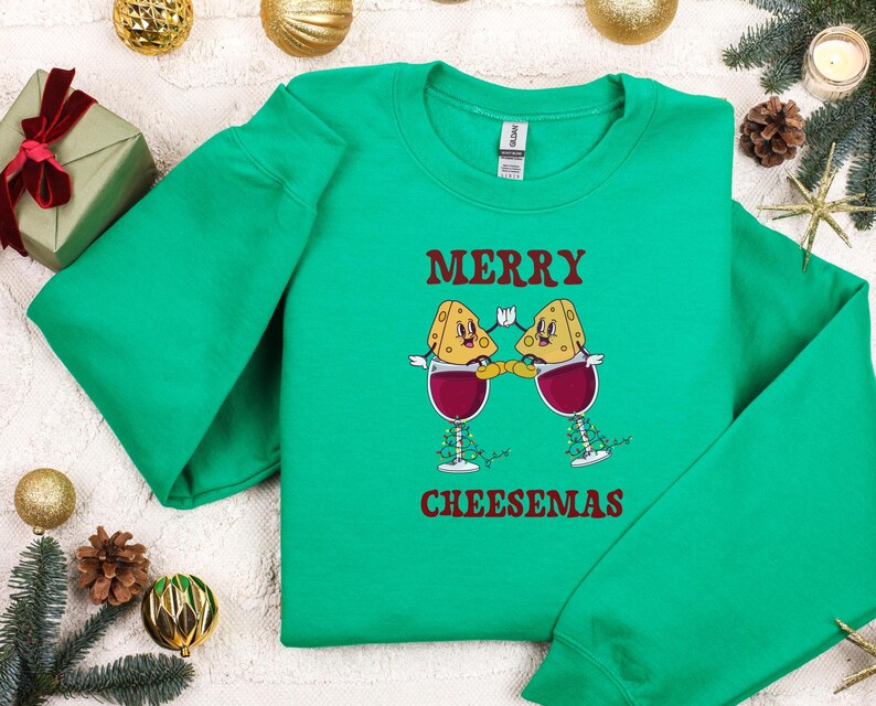 May include: Green Christmas sweater with a festive design featuring two cheese wedges wearing Santa hats and holding glasses of wine. The text "Merry Cheesemas" is printed on the sweater.
