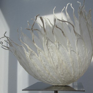 Könnte beinhalten: Eine weiße Papierskulptur, die einer Blume mit zarten, fließenden Blütenblättern ähnelt. Die Skulptur wird auf einem silbernen Ständer präsentiert.