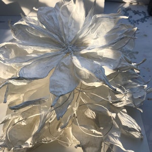 May include: A large, handmade white paper flower with multiple layers of petals. The flower has a detailed centre and delicate, textured petals. The lighting creates shadows, highlighting the flower's form and texture. The overall effect is elegant and artistic.