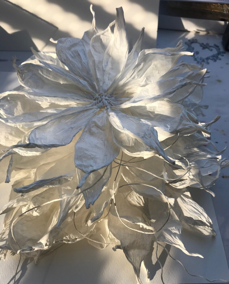 May include: A large, handmade white paper flower with multiple layers of petals. The flower has a detailed centre and delicate, textured petals. The lighting creates shadows, highlighting the flower's form and texture. The overall effect is elegant and artistic.