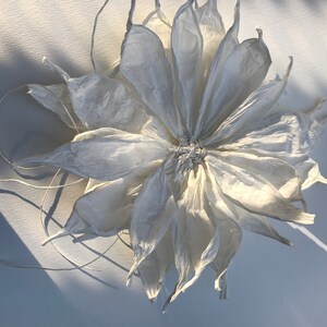 May include: A large, white paper flower with multiple layers of delicate petals. The flower's centre is tightly woven, and thin, white strings extend from it. The petals have a textured, crinkled appearance, and the flower casts a soft shadow.