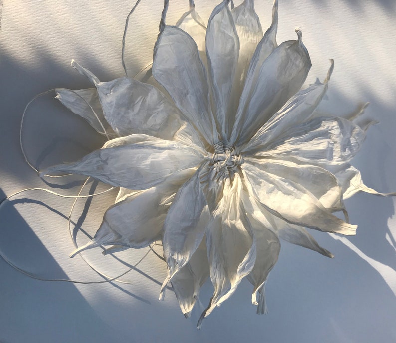 May include: A large, white paper flower with multiple layers of delicate petals. The flower's centre is tightly woven, and thin, white strings extend from it. The petals have a textured, crinkled appearance, and the flower casts a soft shadow.