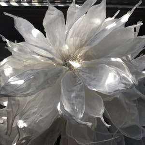 May include: A large, white, paper flower with multiple layers of delicate petals. The flower is illuminated, creating highlights and shadows that emphasise its texture and form. The overall effect is soft and ethereal.
