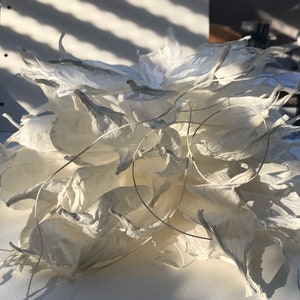 May include: A white paper sculpture, floral in appearance, with multiple layers of petal-like shapes. Fine wire details are visible. The sculpture is illuminated by sunlight, creating shadows and emphasizing the textures. The piece is likely handmade.