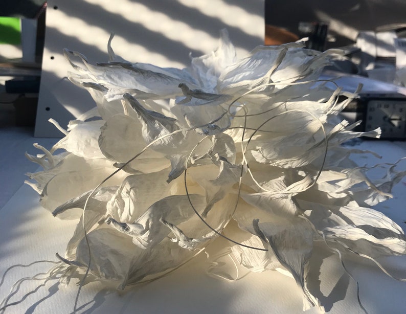 May include: A white paper sculpture, floral in appearance, with multiple layers of petal-like shapes. Fine wire details are visible. The sculpture is illuminated by sunlight, creating shadows and emphasizing the textures. The piece is likely handmade.