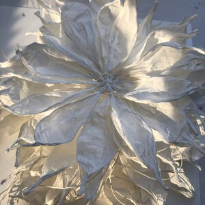 May include: A large, handmade white paper flower with multiple layers of delicate petals. The flower's centre is intricately woven, and the petals have a textured, almost translucent quality. The lighting creates shadows, enhancing the flower's three-dimensional form.