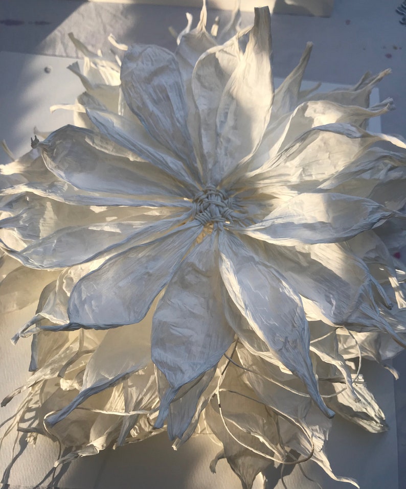 May include: A large, handmade white paper flower with multiple layers of delicate petals. The flower's centre is intricately woven, and the petals have a textured, almost translucent quality. The lighting creates shadows, enhancing the flower's three-dimensional form.