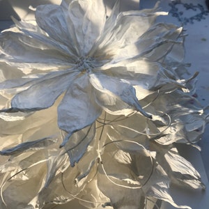 May include: A large, handmade paper flower with multiple layers of white petals. The flower has a detailed, textured appearance, with delicate edges and a central core. The petals are arranged in a layered pattern, creating a full, open bloom. The flower is likely for home decor.