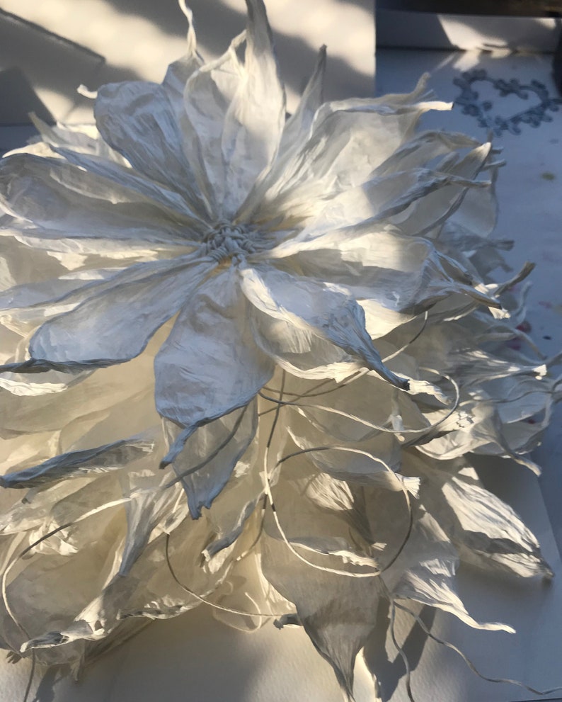 May include: A large, handmade paper flower with multiple layers of white petals. The flower has a detailed, textured appearance, with delicate edges and a central core. The petals are arranged in a layered pattern, creating a full, open bloom. The flower is likely for home decor.