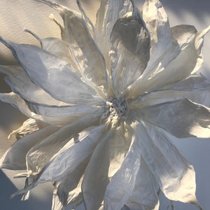 May include: A large, handcrafted paper flower with multiple layers of delicate, off-white petals. The flower's centre is tightly clustered, and the petals fan out, creating a full, textured appearance. The lighting highlights the subtle variations in the paper's surface.