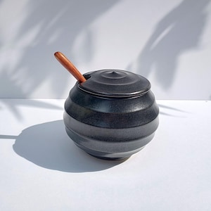 Könnte beinhalten: Eine schwarze Keramik-Zuckerdose mit Deckel und einem Holzlöffel. Die Schüssel hat eine runde Form mit horizontalen Rillen. Der Löffel ist in die Schüssel gesteckt. Der Hintergrund ist weiß mit Schatten.