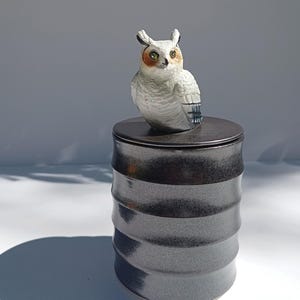 Könnte beinhalten: Ein zylindrischer Keramikbehälter mit einer dunklen, glänzenden, metallisch wirkenden Oberfläche und horizontalen Rillen. Der Deckel ist mit einer detaillierten, handbemalten Figur einer weißen Eule mit orangefarbenen Akzenten verziert.