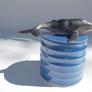 Könnte beinhalten: Ein blauer Keramiktopf mit Deckel, auf dem eine Walfigur steht. Der Topf hat ein geripptes Design und eine glänzende Oberfläche. Der Wal ist dunkelgrau und scheint aus Kunststoff zu sein. Der Schatten des Topfes ist sichtbar.