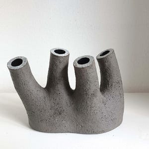 Könnte beinhalten: Eine dunkelgraue, mehrstielige Keramikvase mit rauer, texturierter Oberfläche. Das Objekt verfügt über vier ausgeprägte, nach oben gerichtete zylindrische Öffnungen, die in einer Reihe angeordnet sind, jede mit einem hohlen Zentrum und einem dunklen Innenbereich.