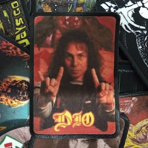 May include: A black and red embroidered patch featuring a man with long hair and a black shirt. The man is making the rock and roll hand gesture. The text "DIO" is written in yellow on the patch.