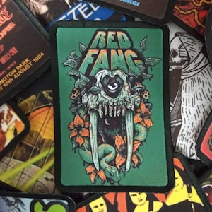 May include: A green embroidered patch with the text "RED FANG" in white. The patch features a skull with a single eye and a large tusk, surrounded by flowers and a snake.