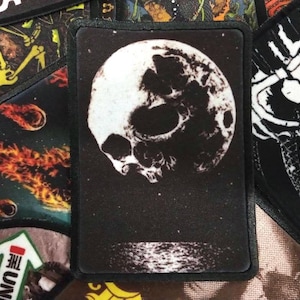 May include: Black and white embroidered patch featuring a skull on a moon. The patch has a black border and is sewn onto a black fabric background.
