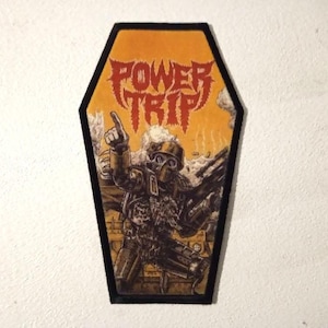 Könnte beinhalten: Ein schwarzer und gelber Sarg-förmiger Patch mit dem Bandnamen "POWER TRIP" in roten Buchstaben. Der Patch zeigt einen Soldaten mit Gasmaske und Waffe, der vor einem Panzer steht.