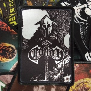 May include: Black and white embroidered patch with a hooded figure holding a sword. The figure is standing in front of a swirling background. The word "VOMIT" is written in a gothic font below the figure.