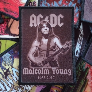 Puede incluir: Un parche en blanco y negro con una foto de Malcolm Young, guitarrista de la banda AC/DC, tocando la guitarra. El texto "AC/DC" está por encima de la foto y "Malcolm Young" está por debajo de la foto. Los años "1953-2017" están debajo del nombre.