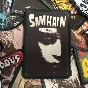 May include: A black and white patch featuring the band name "SAMHAIN" above a stylized face. The patch is rectangular with a black border, surrounded by other patches with various designs and text.